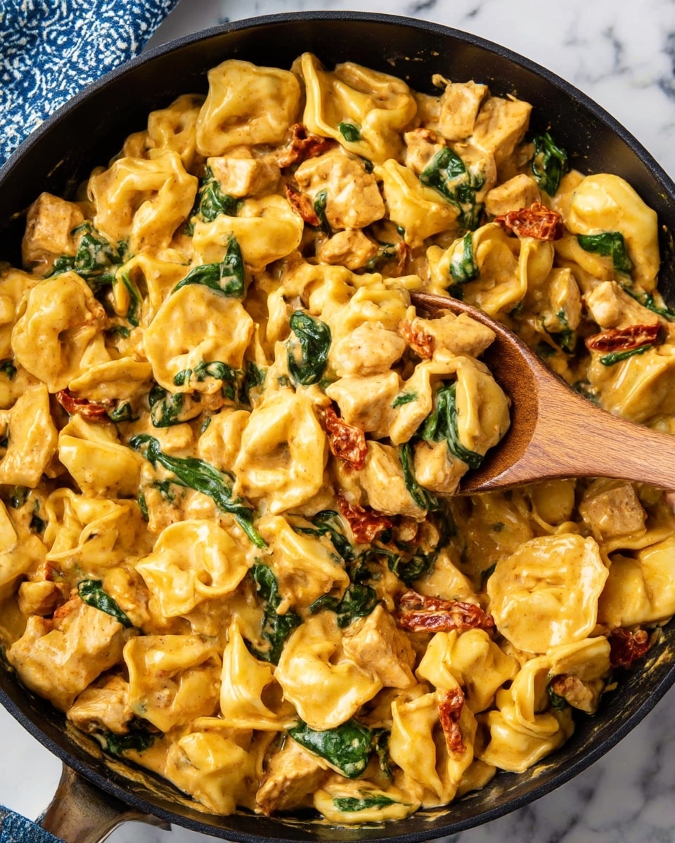 A black pan filled with tortellini pasta in a creamy orange-yellow sauce mixed with green spinach leaves and light brown chicken pieces, with some red sun-dried tomato bits scattered throughout. A wooden spoon rests in the pan, partially covered with the sauce and pasta. The pan sits on a white marbled surface with a small part of a blue and white pattern cloth visible at the top left corner. photo taken with an iphone --ar 4:5 --v 7