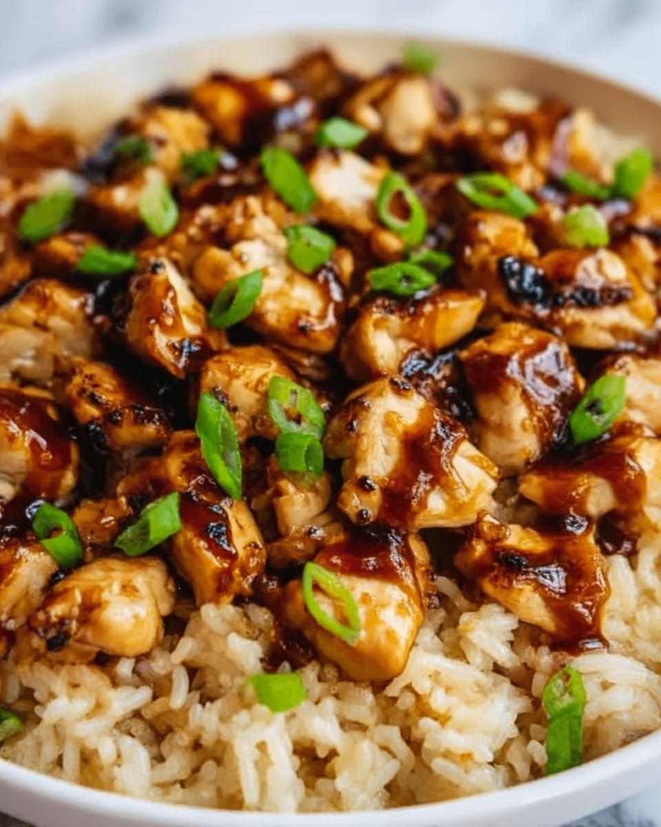 The image shows a close-up of a dish with two visible layers: the bottom layer is light brown, cooked rice with a fluffy texture, and the top layer consists of bite-sized pieces of grilled chicken coated in a shiny, dark brown sauce. Small green onion slices are sprinkled on the chicken, adding a fresh splash of color. The overall look is rich and glossy, with the rice and chicken clearly separated. The background is a white marbled texture. Photo taken with an iphone --ar 4:5 --v 7