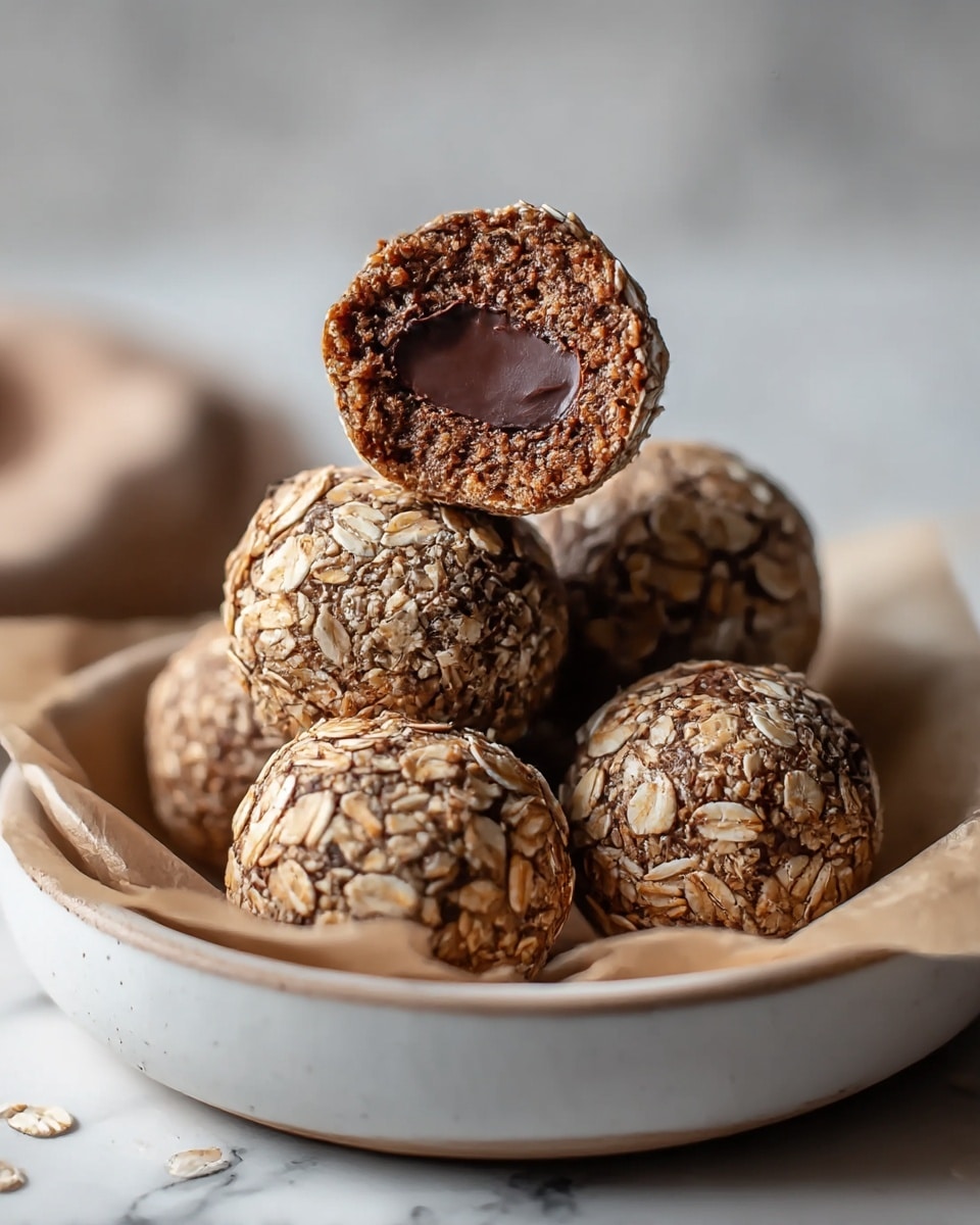 The image shows four round chocolate oat balls resting on a piece of parchment paper inside a shallow white ceramic bowl. The oat balls have a rough outer layer made of large, light brown oat flakes. One oat ball is cut in half and placed on top of the others, showing a dense, dark brown chocolate interior with a smooth, creamy chocolate layer in the center. The background is softly blurred with a white marbled surface visible. photo taken with an iphone --ar 4:5 --v 7