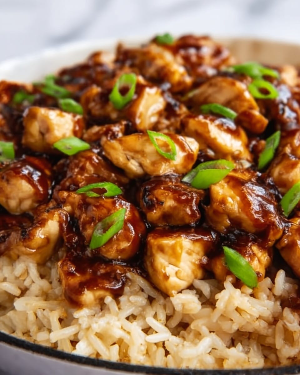 The image shows a close-up of a dish with two main layers. The bottom layer is light brown cooked rice, with fluffy and soft texture. On top, there are pieces of grilled chicken that are golden brown with some darker grill marks, covered in a shiny, dark brown sauce. Small, round green onion pieces are scattered on top, adding a fresh green contrast to the warm colors. The dish is on a white plate, set on a white marbled surface. photo taken with an iphone --ar 4:5 --v 7