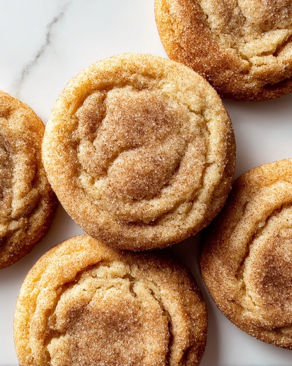 The image shows five soft cookies arranged closely on a white marbled surface. Each cookie is round with a slightly cracked texture, featuring light brown dough with a darker brown cinnamon sugar coating on top. The tops of the cookies have a sugary sparkle, giving a granular texture visible across their surfaces. The edges of the cookies are slightly raised with a crinkled look, and the centers are more indented and smoother. The warm color tones of the cookies contrast nicely with the clean, white marbled background. photo taken with an iphone --ar 4:5 --v 7