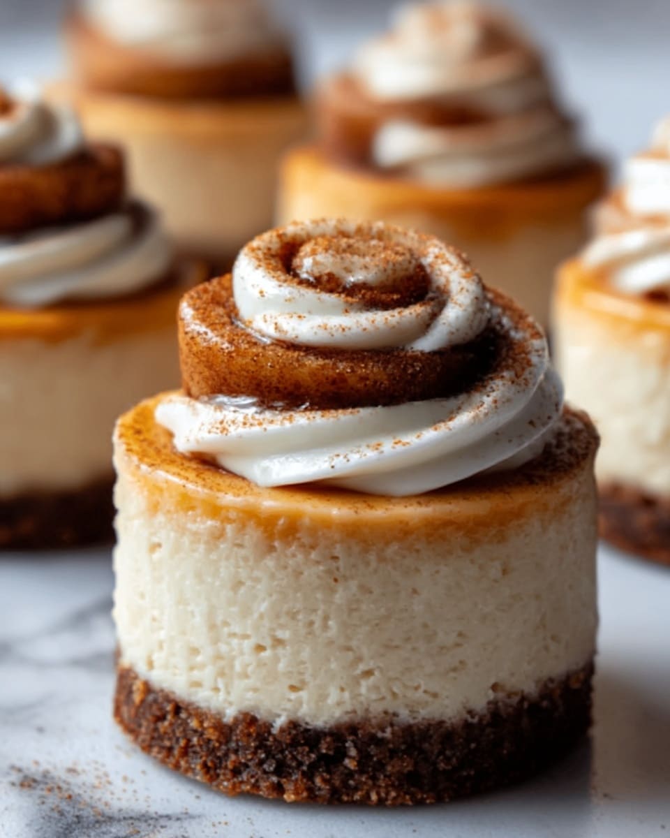 The image shows a small round dessert with three visible layers set on a white marbled surface. The bottom layer is a dark brown crust that looks crumbly. Above it is a creamy beige layer, smooth and thick. On top sits a swirl of cinnamon roll dough, golden brown with a soft texture, dusted slightly with cinnamon. The cinnamon roll is decorated with a shiny white frosting swirl that contrasts with the dough’s rich color. In the background, more of these desserts are softly out of focus. Photo taken with an iphone --ar 4:5 --v 7