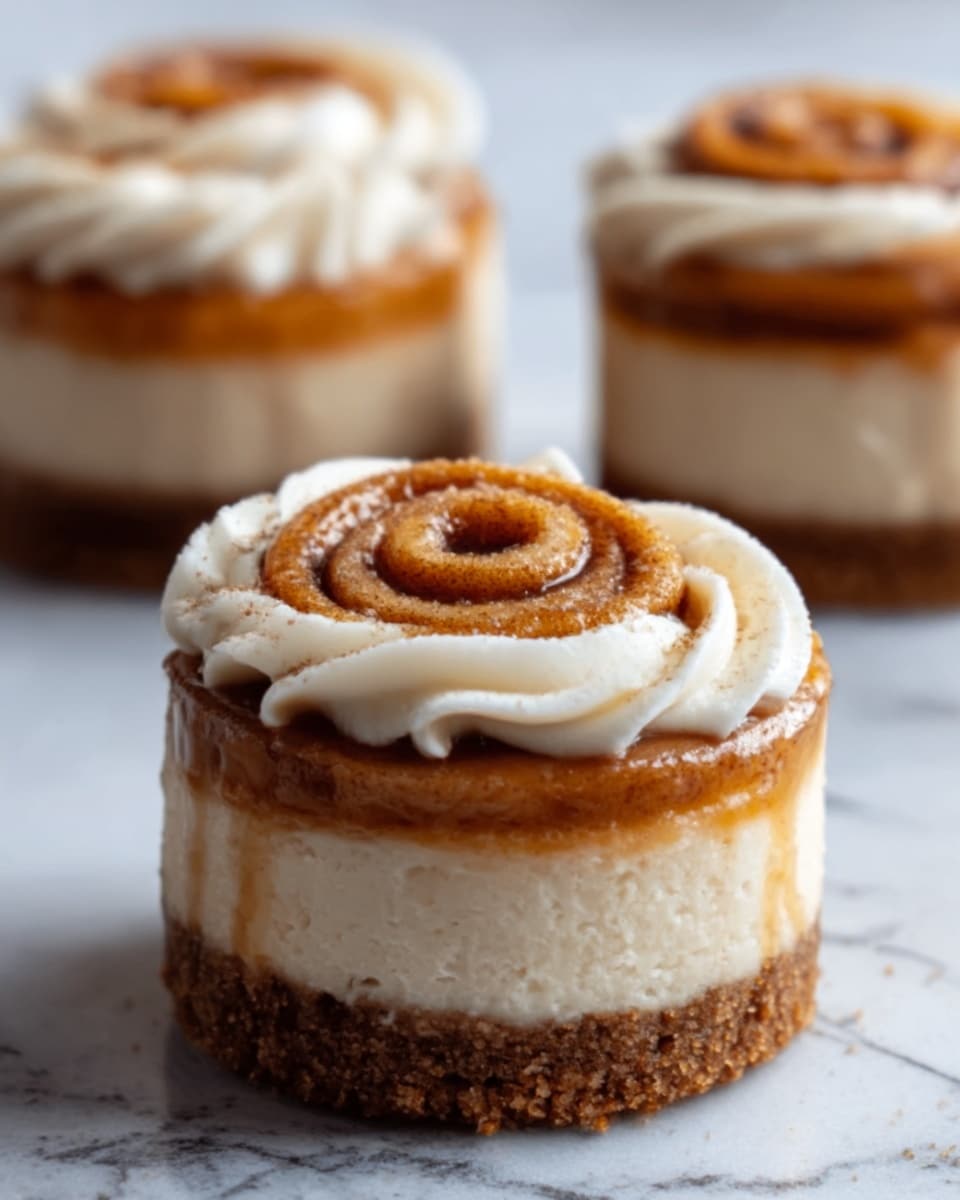 The image shows a close-up of a small dessert shaped like a cinnamon roll on a white marbled surface. The dessert has three clear layers: a bottom crumbly brown crust, a thick middle creamy beige layer, and a top swirled cinnamon roll with a golden-brown color. White cream frosting is piped around the cinnamon roll in a spiral pattern, adding texture and contrast to the dessert. In the background, two more similar desserts are slightly blurred, sitting on the same white marbled surface. Photo taken with an iphone --ar 4:5 --v 7