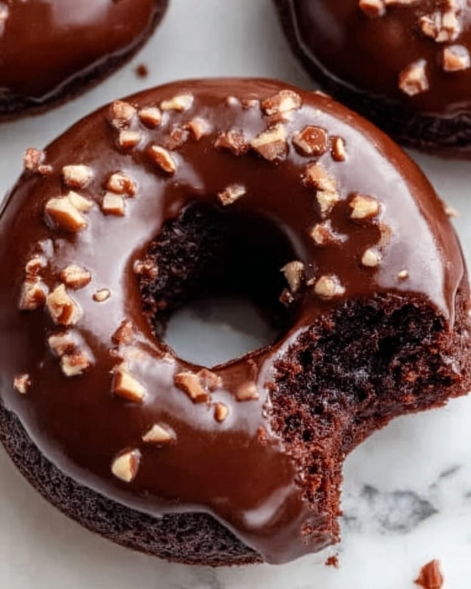 The image shows a close-up of a glossy chocolate donut with a shiny, smooth chocolate glaze covering the top. Small pieces of chopped nuts are sprinkled on the glaze, adding texture. One donut has a bite taken out, revealing a dark, moist, and soft chocolate cake inside with a fluffy texture. The background is a white marbled surface. photo taken with an iphone --ar 4:5 --v 7