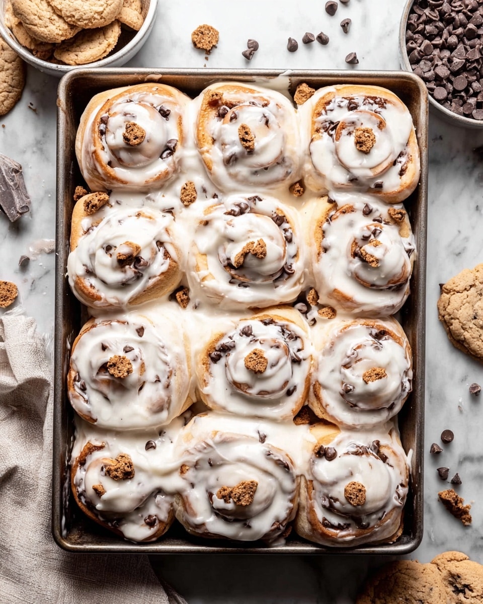 A metal baking tray filled with ten rolled cinnamon buns covered in thick white icing that softly drips over the edges. The buns are light brown with dark chocolate chips visible in the swirls. Small, round mini chocolate chip cookies and cookie crumbs are scattered on top of the icing and around the buns, adding texture. The tray is placed on a white marbled surface with broken cookie pieces, chocolate chips, and a bowl with more mini cookies nearby. A light-colored cloth napkin is to the side, adding softness to the scene. Photo taken with an iphone --ar 4:5 --v 7
