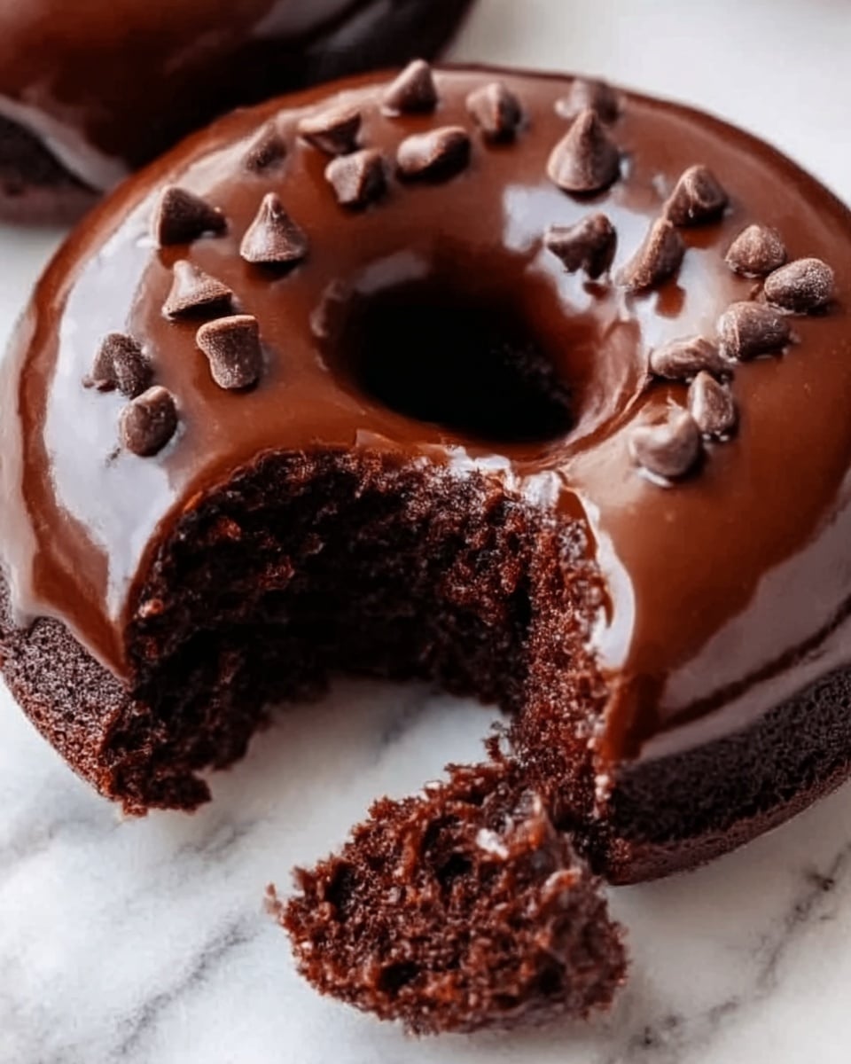 The image shows a close-up of two round chocolate cakes placed on a white marbled surface. Each cake is covered with shiny, smooth chocolate glaze on top, reflecting light softly. The top layer is decorated with small, scattered pieces of chocolate chips. One cake has a bite taken out, revealing three layers inside: the outermost layer is dark, moist chocolate cake, the middle layer is thick, creamy chocolate filling, and the top layer is the glossy chocolate glaze. The texture of the cake looks soft and rich. Photo taken with an iphone --ar 4:5 --v 7