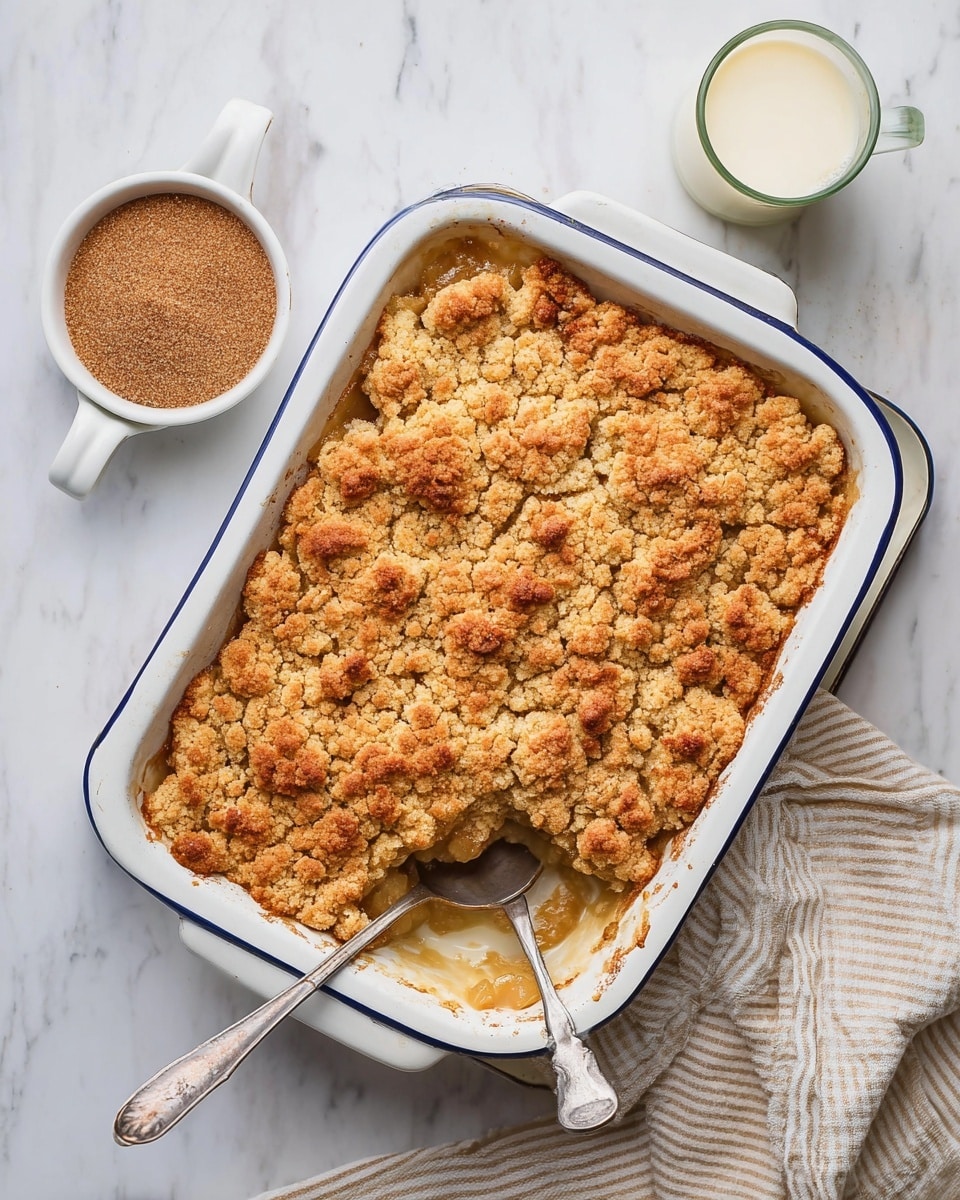 A white baking dish with blue trimming holds a golden brown crumble topped dessert, showing a textured, crumbly layer that is uneven and crispy on top. Inside the dish, part of the dessert has been scooped out with a silver spoon resting inside the hollowed area, revealing a soft, moist layer beneath the crumbly top. To the upper left of the dish is a small white ramekin filled with brown sugar or spice powder, and to the upper right, there is a white glass filled with light cream or milk. Everything is placed on a white marbled surface with a beige and white striped cloth partially visible at the bottom right corner. Photo taken with an iphone --ar 4:5 --v 7