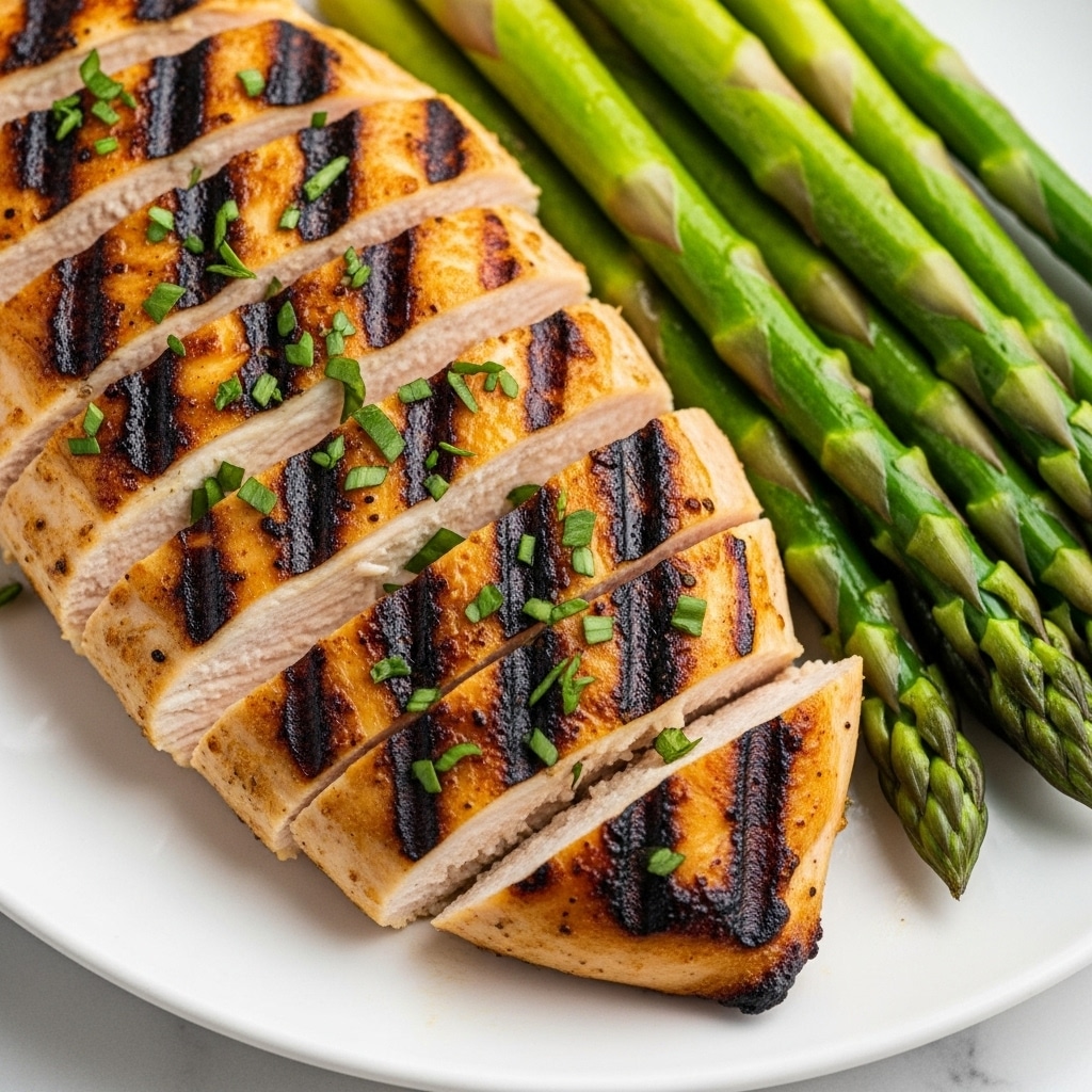 A close-up view of a grilled chicken breast sliced into thick pieces, showing a golden-brown color with dark grill marks across the top. The chicken is garnished with small green herbs sprinkled evenly on top. Alongside the chicken are bright green asparagus spears laying next to the slices, adding contrast to the dish. The food rests on a white plate, all set against a white marbled texture. photo taken with an iphone --ar 4:5 --v 7