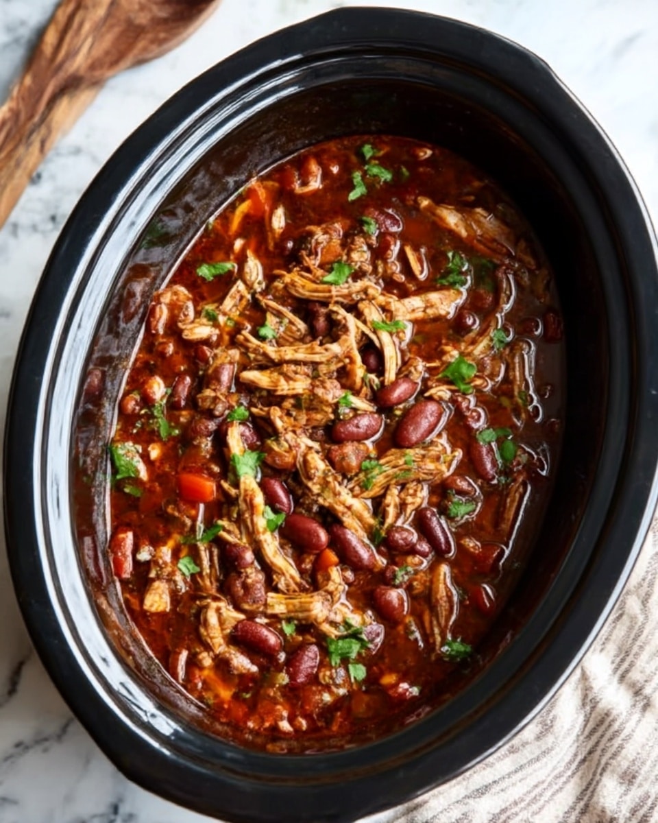 The image shows a black slow cooker filled with a rich, dark red stew. On top, there are shredded pieces of light brown chicken, mixed with small chunks of reddish-brown beans and bright red bell peppers. Tiny green parsley leaves are sprinkled over the dish, adding a fresh touch. The slow cooker sits on a white marbled surface with a striped cloth nearby and some wooden utensils in the background. The textures are moist and tender, with a shiny, thick broth surrounding the ingredients. photo taken with an iphone --ar 4:5 --v 7