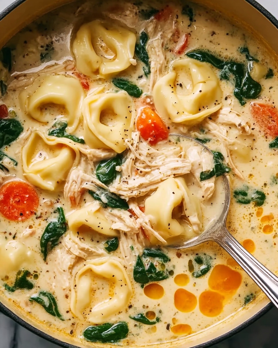 This close-up image shows a creamy soup filled with several layers of ingredients. The top layer consists of smooth, light yellow tortellini pasta floating among shredded white and light brown chicken pieces. Bright orange carrot slices and dark green spinach leaves are spread throughout the soup, adding color contrast. The creamy broth is light beige with specks of black pepper and some orange oil droplets floating on the surface. A silver spoon is partially dipped into the soup on the right side. The pot containing the soup is placed on a white marbled texture. Photo taken with an iphone --ar 4:5 --v 7