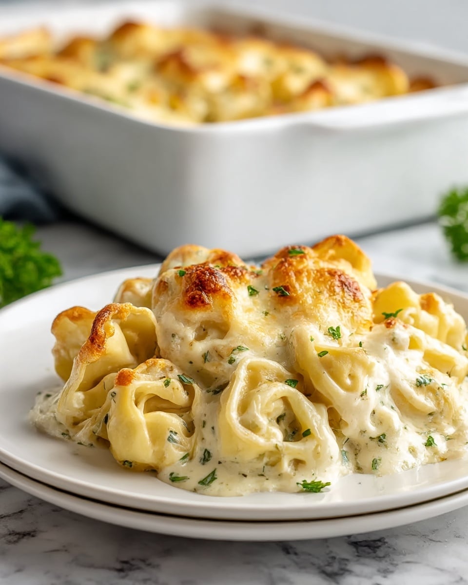 A white plate holds a serving of baked tortellini pasta covered with a creamy white cheese sauce. The pasta is folded into small rounds with visible folds and is topped with a bubbly, golden-brown melted cheese layer. Small green herb bits are sprinkled on top and throughout the dish, adding specks of color to the creamy texture. In the background, a white baking dish with more of the same pasta is slightly out of focus on a white marbled surface. photo taken with an iphone --ar 4:5 --v 7