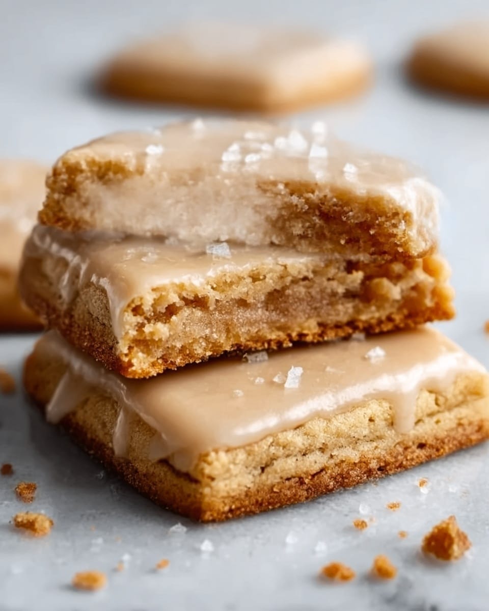 The image shows a close-up of two square-shaped cookies stacked on a white marbled surface. The top cookie is broken in half, revealing a soft, brown, crumbly inside layer with a slightly gooey texture. Both cookies are covered with a light tan glaze that glistens under the light, with some small white salt crystals sprinkled on top. The edges of the cookies are slightly crinkled and golden brown, giving a crispy look. Crumbs are scattered around, adding detail to the scene. photo taken with an iphone --ar 4:5 --v 7
