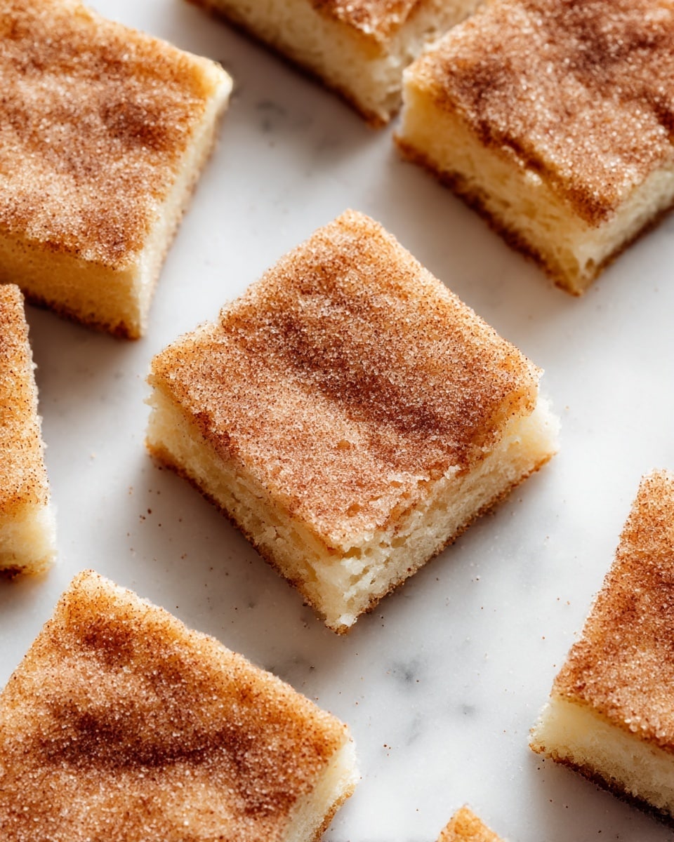 Squares of soft, light beige cake are cut into even pieces and arranged on a white marbled surface. Each piece has a fine, grainy layer of cinnamon and sugar dusted thickly on top, giving a speckled brown and white texture that looks slightly grainy and crunchy. The cake’s crumb shows a soft, moist inside with tiny holes, and the edges are clean but soft. The top surface of each square has light folds and slight dimples, adding a natural, homemade look. photo taken with an iphone --ar 4:5 --v 7