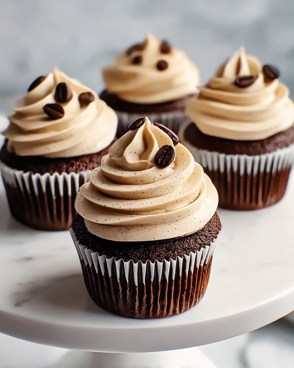 The image shows four chocolate cupcakes on a white round cake stand with a smooth surface. Each cupcake has one dark brown cake layer with a moist and slightly crumbly texture. On top of each cake layer is a thick swirl of light beige coffee-colored frosting, creamy and smooth with small specks visible. The frosting is piped in a tall spiral shape, rising from the edge toward the center, and the top is decorated with several dark brown coffee beans scattered for garnish. The background and surface are white with a marbled texture, giving a clean and bright look. photo taken with an iphone --ar 4:5 --v 7