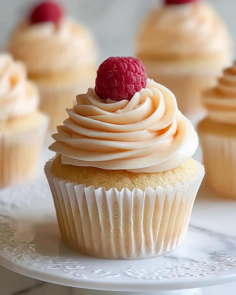 A close-up of a vanilla cupcake with one layer of light golden cake topped with a thick swirl of smooth, creamy pale peach frosting. On top of the frosting sits a single frosty red raspberry. The cupcake is wrapped in a white paper liner and placed on a white plate with delicate embossed patterns, all set against a white marbled surface. In the background, similar cupcakes create a soft, blurred effect. photo taken with an iphone --ar 4:5 --v 7