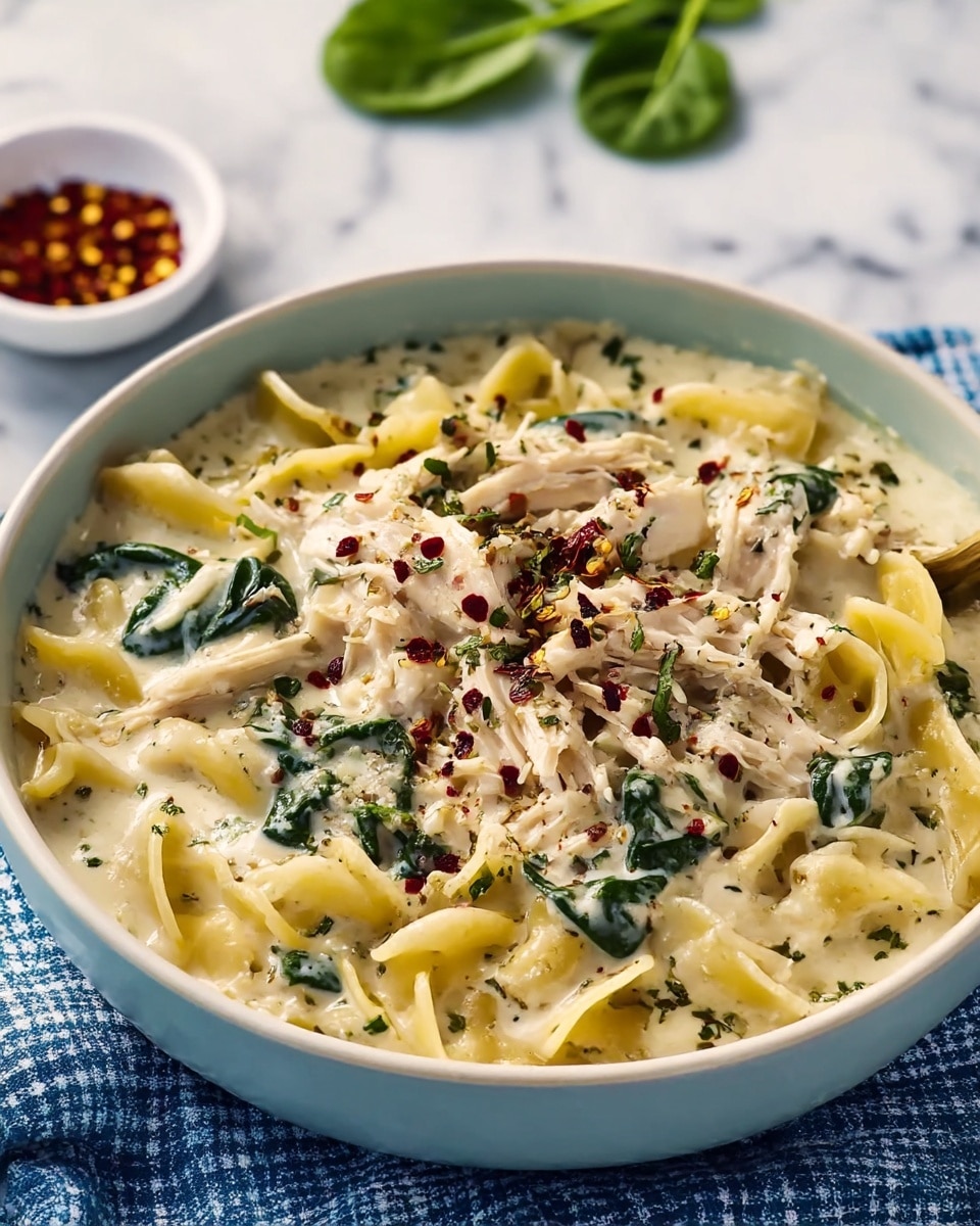 A creamy pasta dish served in a white bowl filled with three main layers: a base layer of tender creamy white sauce, a middle layer of soft, light yellow twisted pasta, and an upper layer of shredded chicken and fresh green spinach leaves mixed throughout, all topped with finely chopped herbs and a sprinkle of dark red chili flakes and black pepper. The bowl rests on a blue-white checkered cloth on a white marbled surface, with a blurred small white bowl of red chili flakes and green leaves visible in the background. Photo taken with an iphone --ar 4:5 --v 7