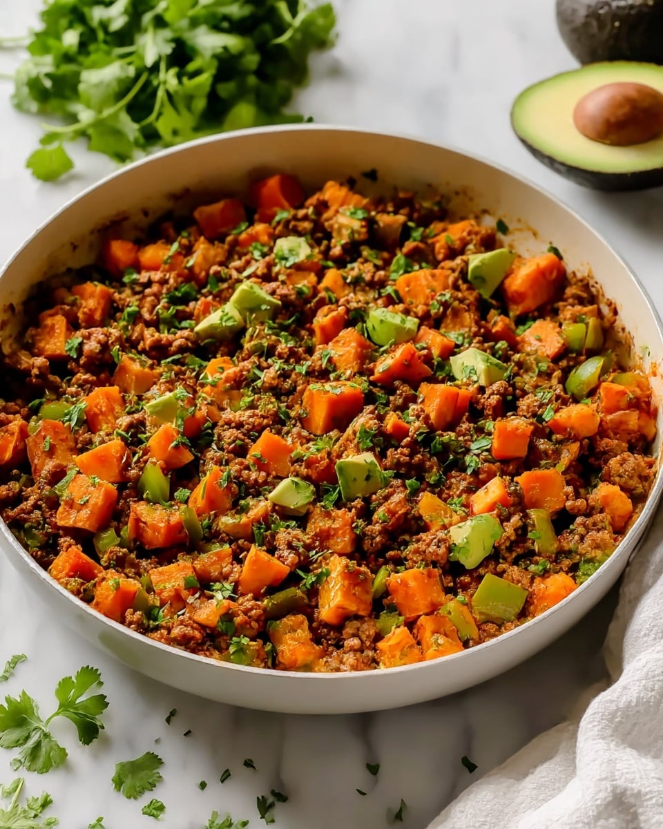 A large white frying pan filled with a cooked mix of small brown ground meat pieces, bright orange diced sweet potatoes, and small chunks of green bell pepper, all garnished with chopped green herbs scattered on top. The pan rests on a white marbled surface with a bunch of green leafy herbs and half of a green avocado in the blurred background. A white towel is partially visible on the right side of the image. photo taken with an iphone --ar 4:5 --v 7