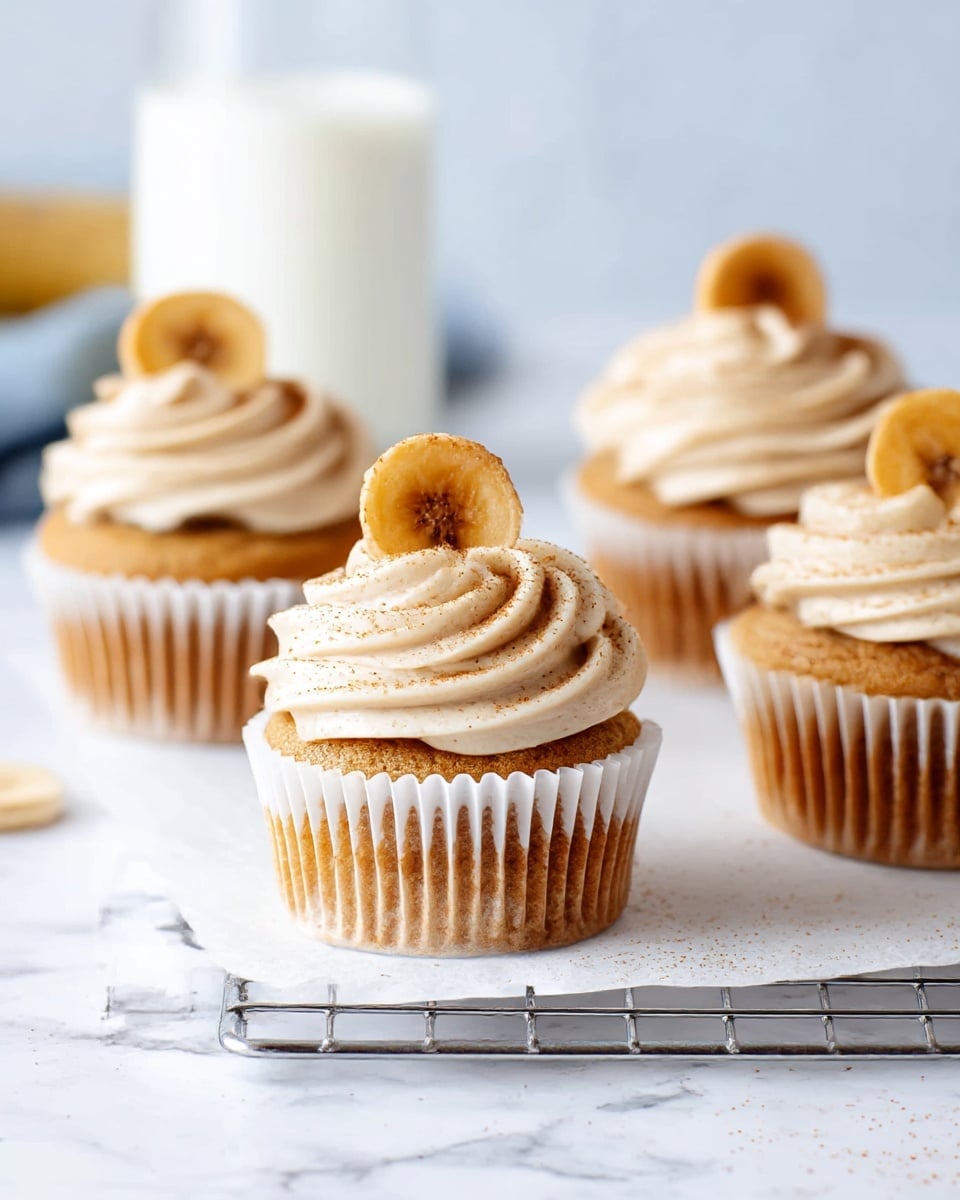 The image shows four light brown cupcakes lined up on a silver wire cooling rack placed over crumpled white paper, all set on a white marbled surface. Each cupcake is topped with a thick swirl of creamy light beige frosting sprinkled lightly with specks of cinnamon. On the top of each swirl, there is a golden-yellow banana chip standing upright. In the blurred background, there is a clear glass of milk against a soft blue backdrop. One banana chip lies next to the cooling rack on the white marbled surface. photo taken with an iphone --ar 4:5 --v 7