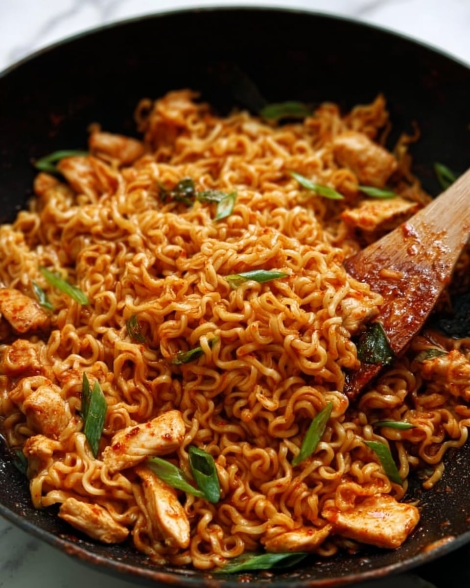 A close-up view of a black pan filled with cooked instant noodles mixed with pieces of cooked chicken and garnished with green onion slices. The noodles are curly and coated in a reddish sauce, making the dish look saucy and flavorful. A wooden spatula lifts some noodles and chicken from the pan, showing the texture of the noodles and meat. The background surface is a white marbled texture. photo taken with an iphone --ar 4:5 --v 7